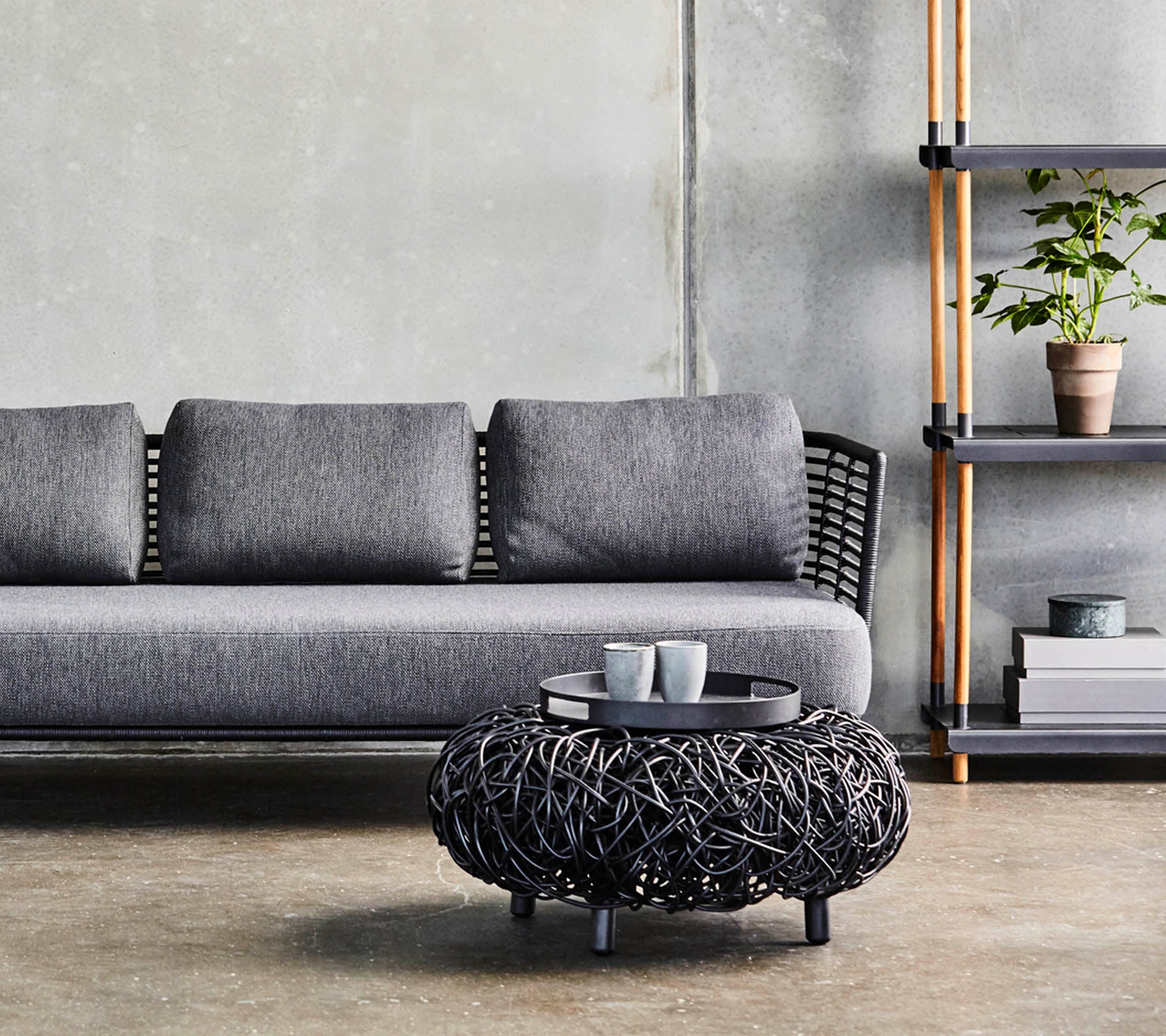 En moderne grå sofa med puter, kombinert med et sirkulært svart flettet kaffebord og en minimalistisk hylle som viser planter og dekor.