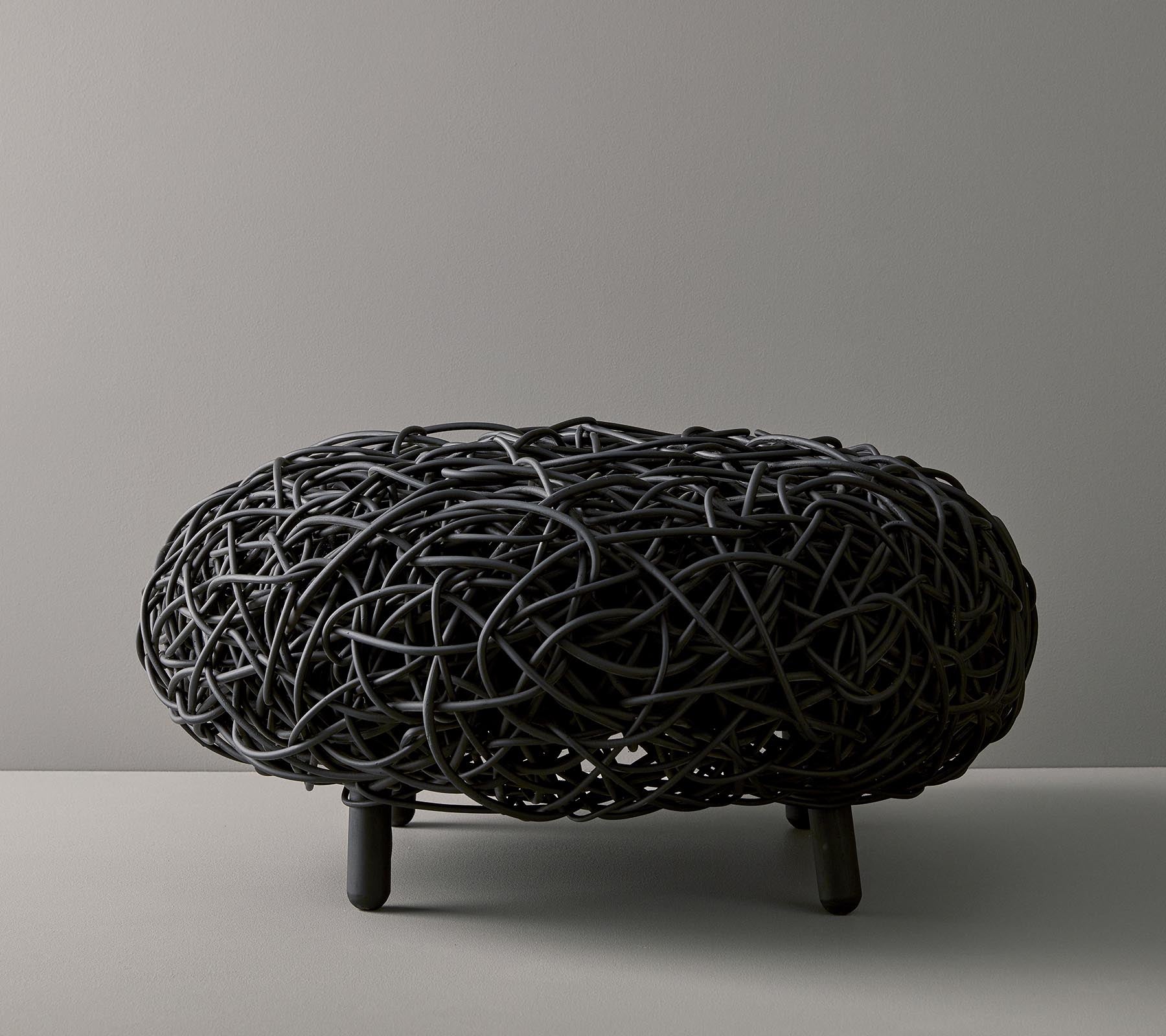 Svart vevd ottoman med en organisk, rund form og intrikat design, hvilende på tre slanke ben mot en nøytral bakgrunn.