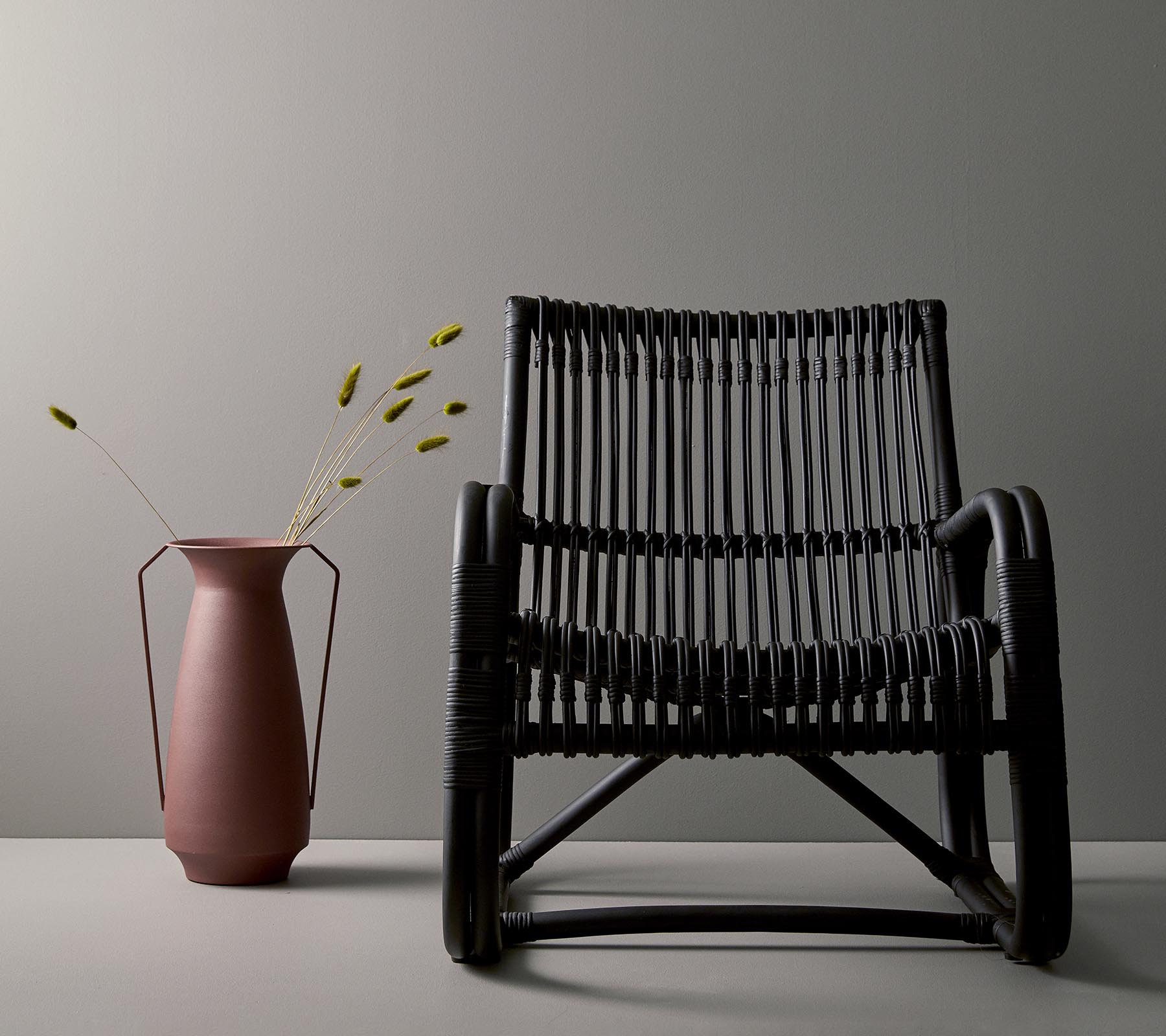 Stilig svart vevd stol ved siden av en rosa vase med grønne stilker, mot en minimalistisk grå bakgrunn.