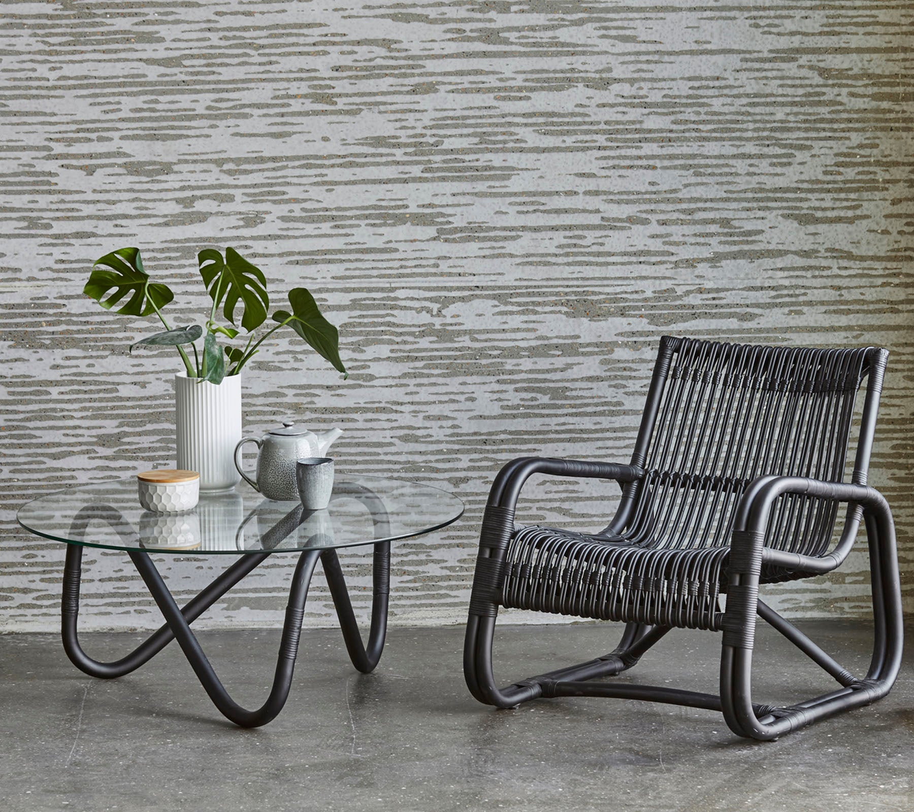 En stilfull stol og et glassbord med en plante og kaffekopper, supplert med en teksturert vegg i bakgrunnen.