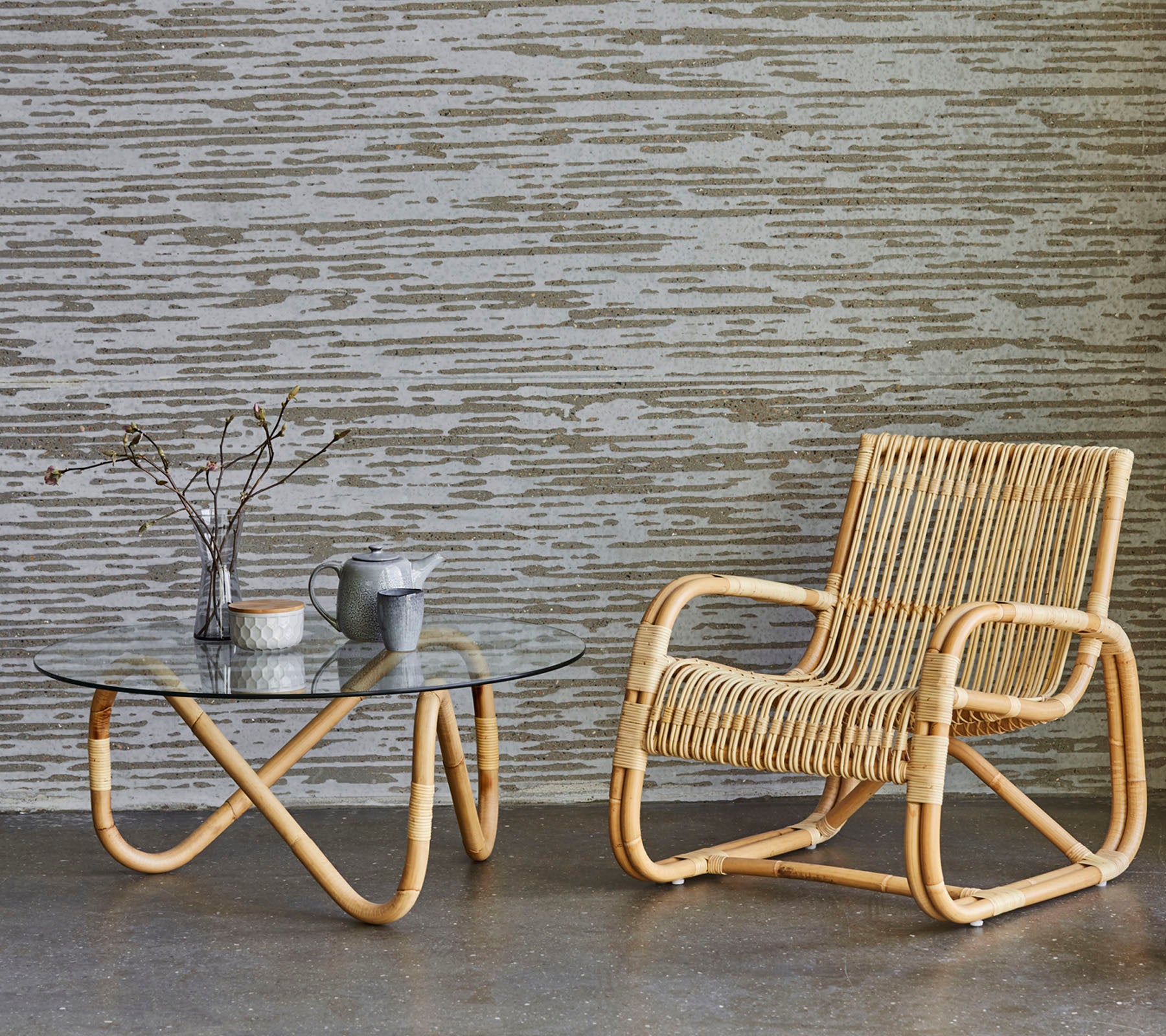 En stilfull rattan stol ved siden av et glass kaffebord, med en vase med grener og en liten tekanne, plassert mot en teksturert bakgrunn.
