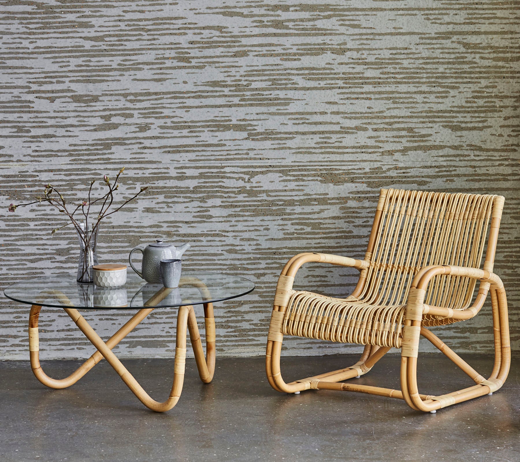 Rattan stol og bord med glasskriveplate med dekorative gjenstander, plassert mot en teksturert vegg. En minimalistisk og moderne dekorasjonsstil.
