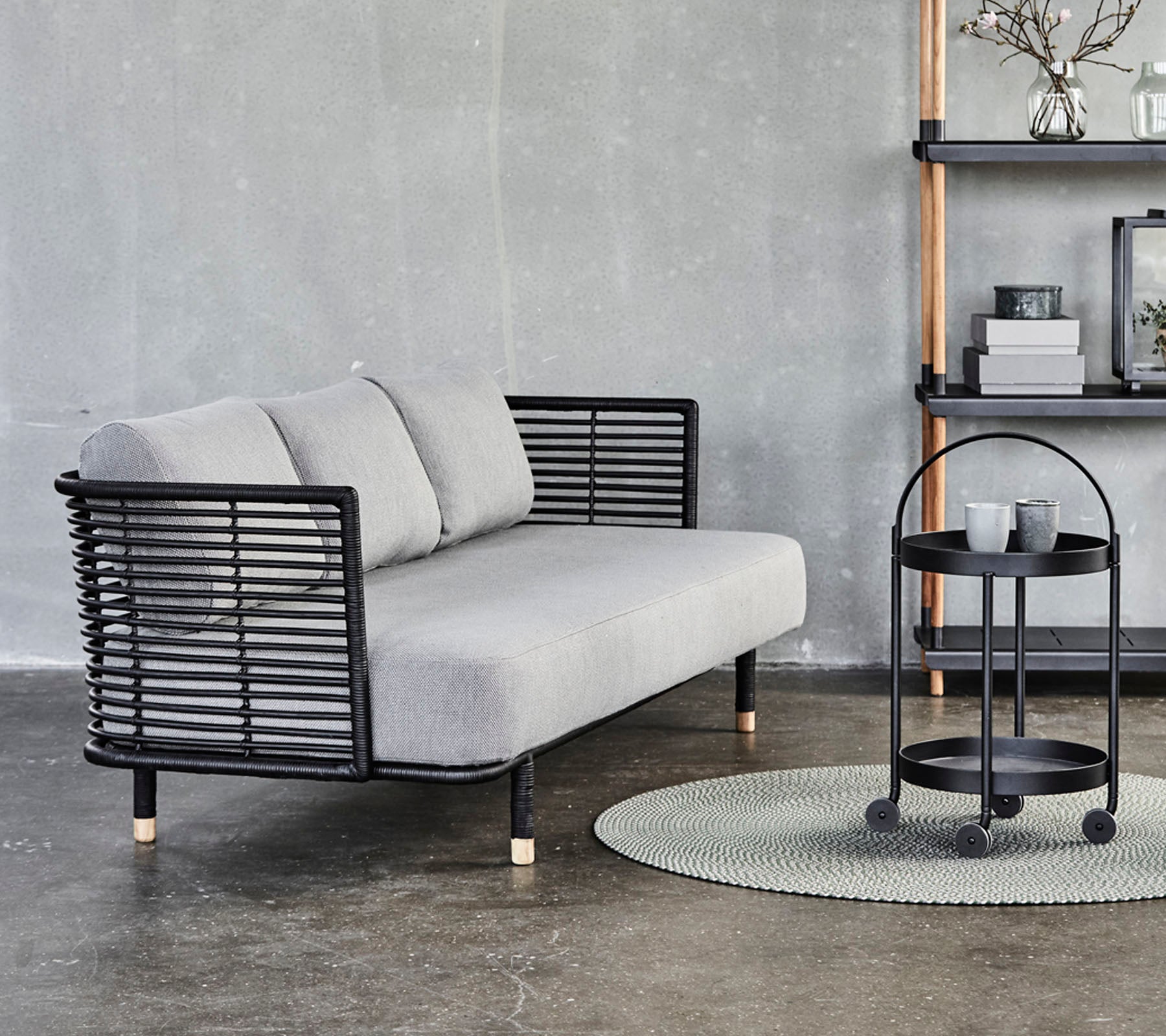 Slank grå sofa med svarte metallakenter og puter, sammen med et rundt sidebord på et teksturert teppe mot en nøytral bakgrunn.