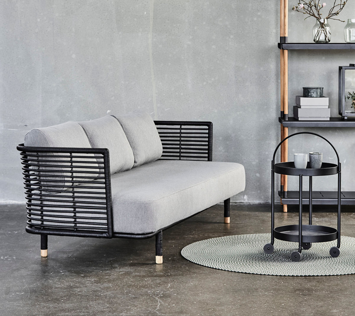 Slank grå sofa med svarte metallakenter og puter, sammen med et rundt sidebord på et teksturert teppe mot en nøytral bakgrunn.