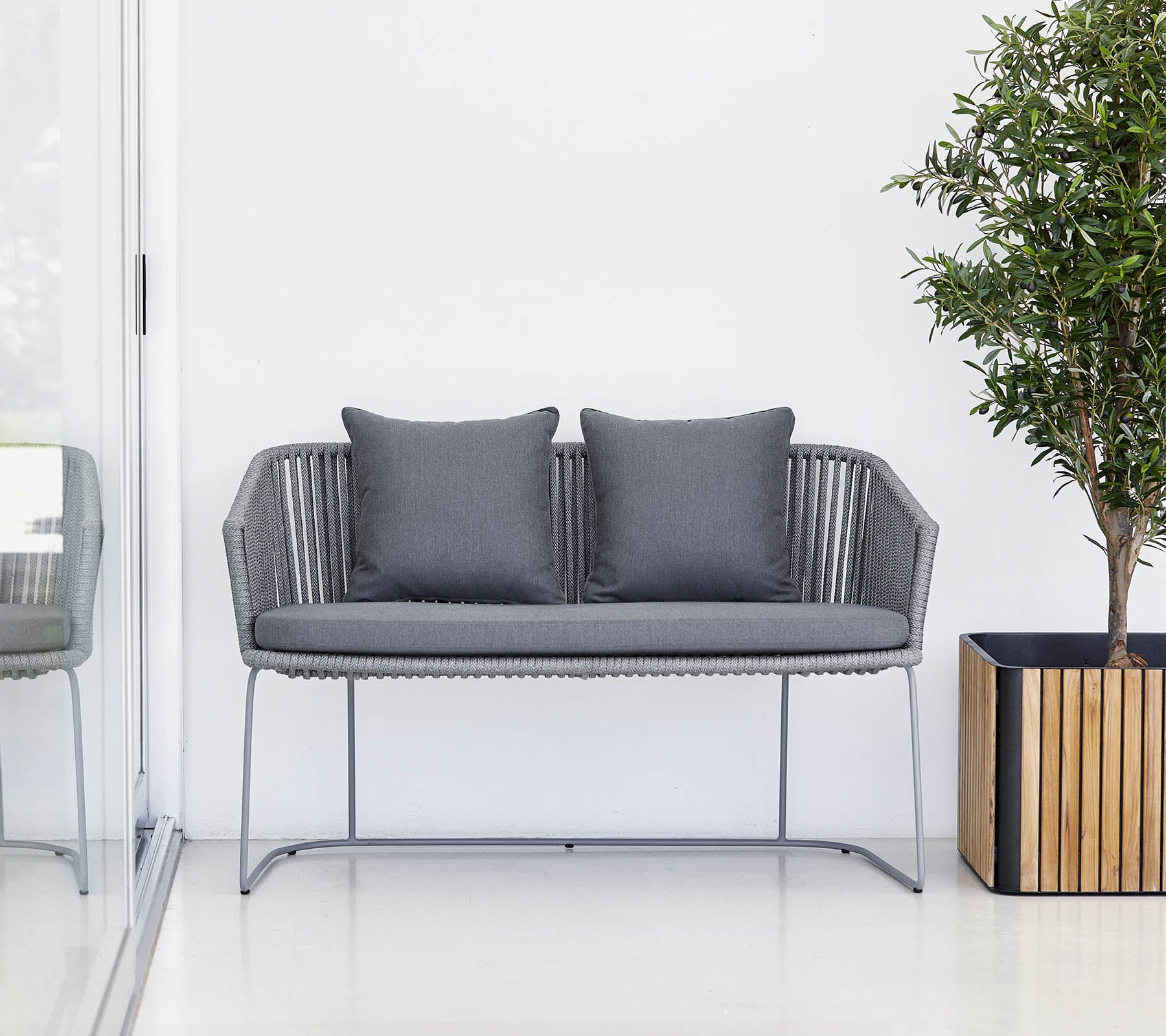 En moderne grå to-seters sofa med puter, akkompagnert av en potteplante og en trekasse.