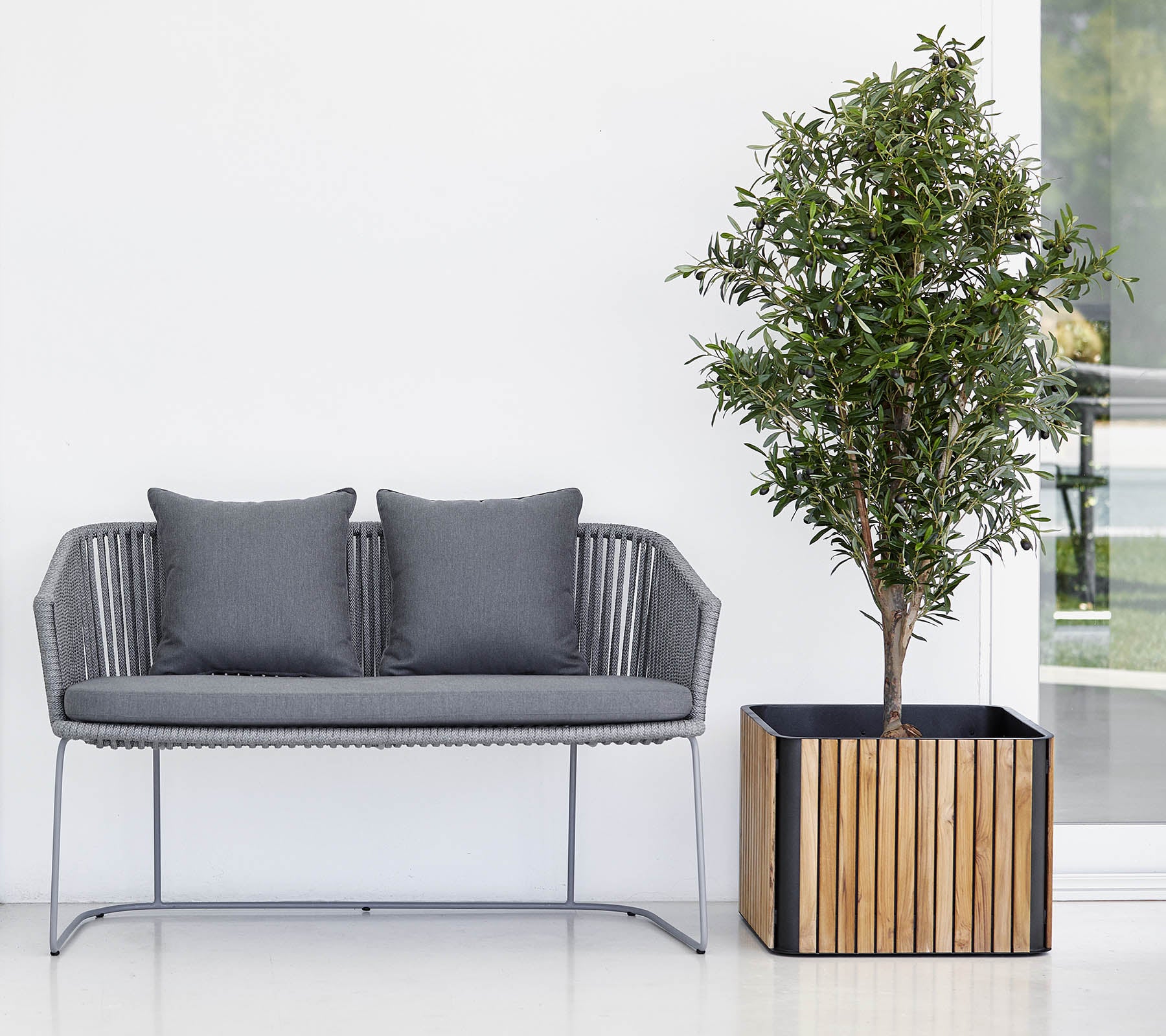 Moderne sofa med to puter ved siden av en potteplante i en dekorativ plantekasse, mot en ren, nøytral bakgrunn.
