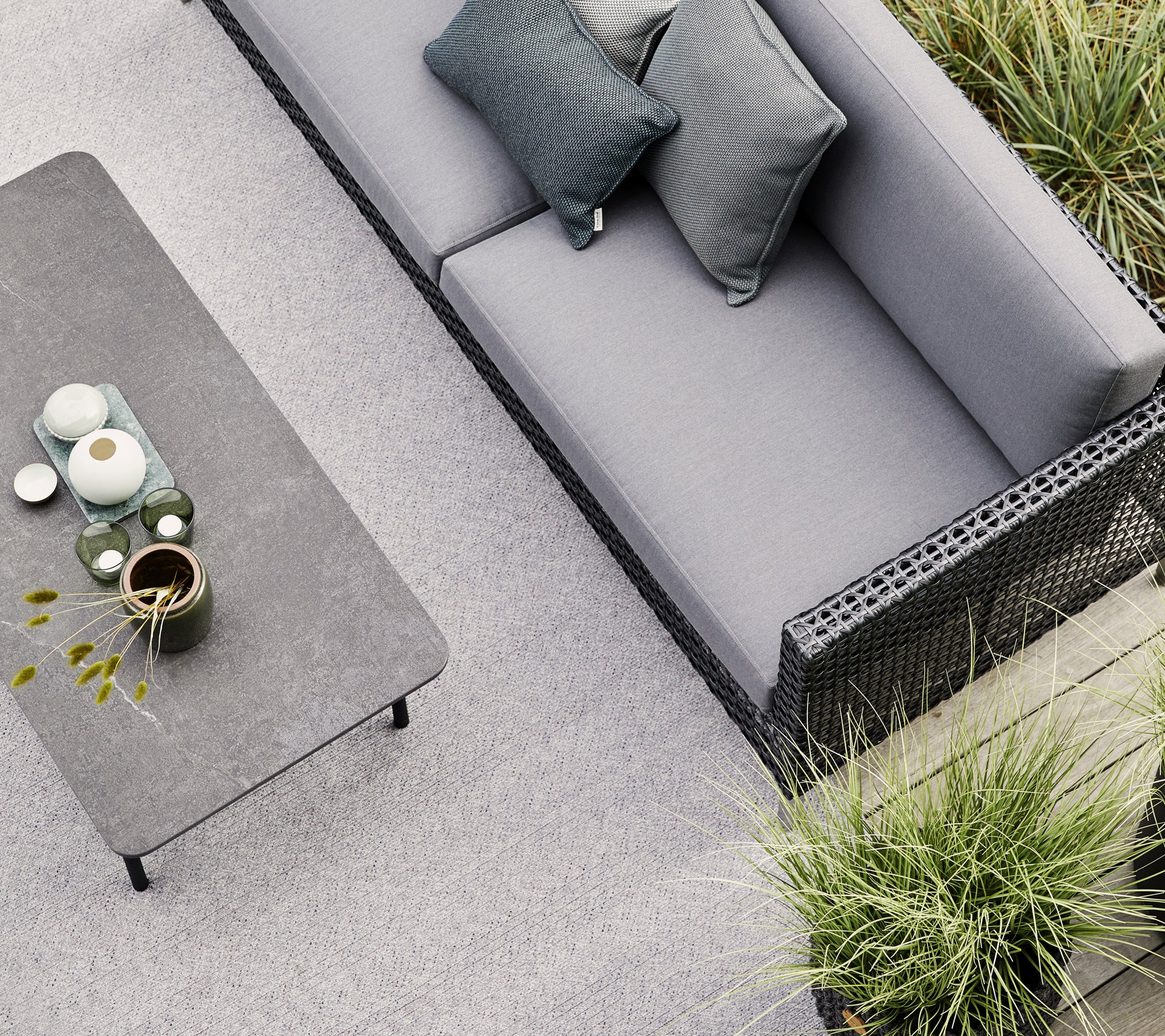 En grå sofa med to puter står ved siden av et minimalistisk bord pyntet med keramikk og en liten plante, omgitt av grønt.