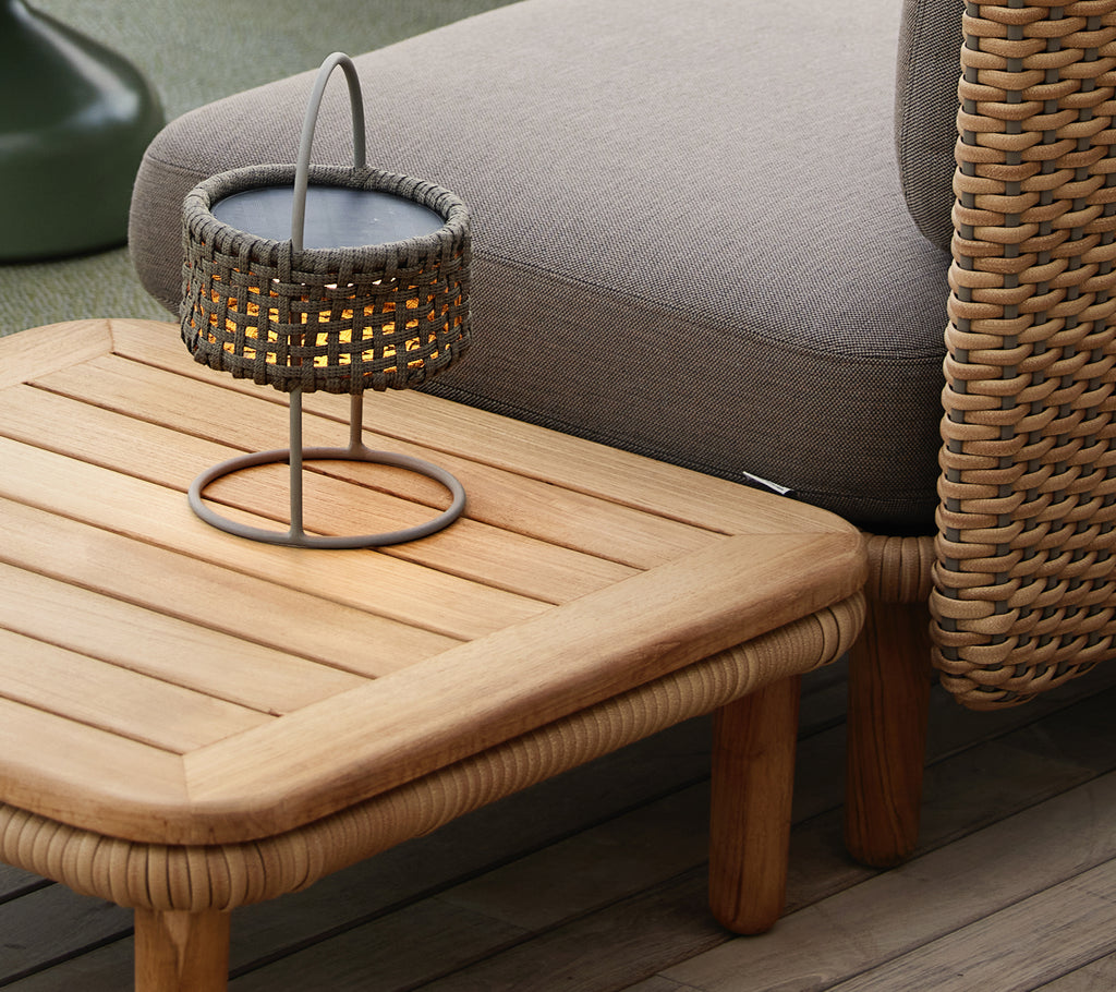 Teak-kaffebord med en lamellert topp, ledsaget av en vevd lampe som skaper en koselig og innbydende atmosfære.