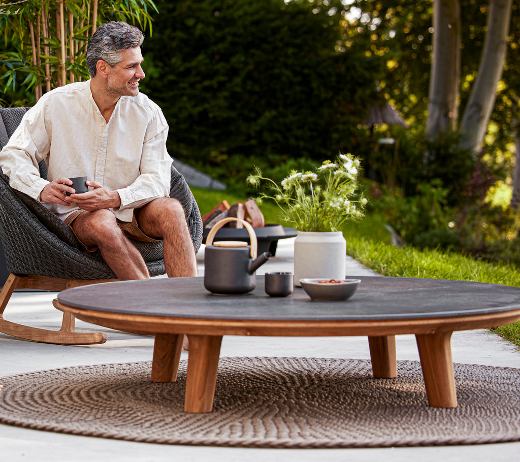 En mann nyter en drink mens han sitter i en gyngestol ved et rundt kaffebord dekorert med stilige serviser og planter.