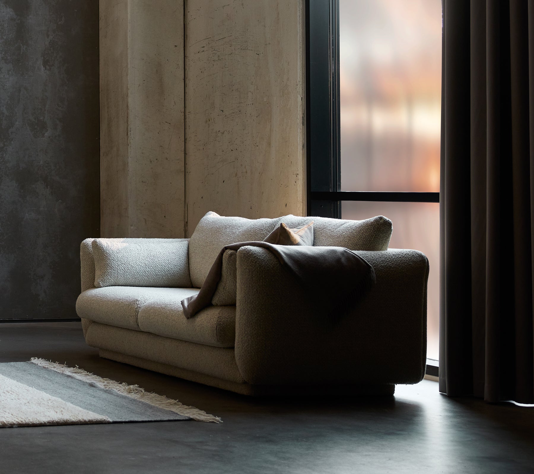 Bloom 2-seter sofa i sandfarge, med en sokkel-design og et koselig teppe drapert over den ene siden.