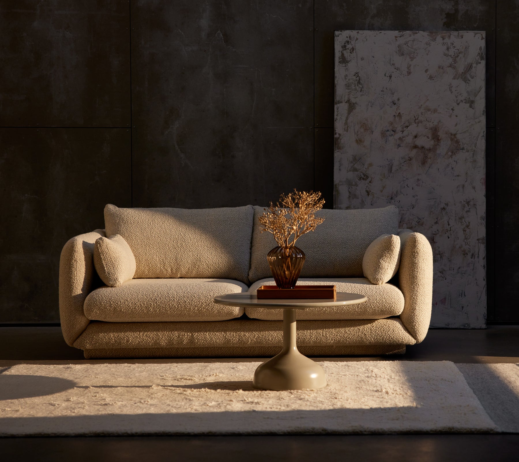 Bloom 2-seters sofa i sand flair med et Glaze kaffebord og en dekorativ plante på et teppe, plassert mot en teksturert mørk vegg.