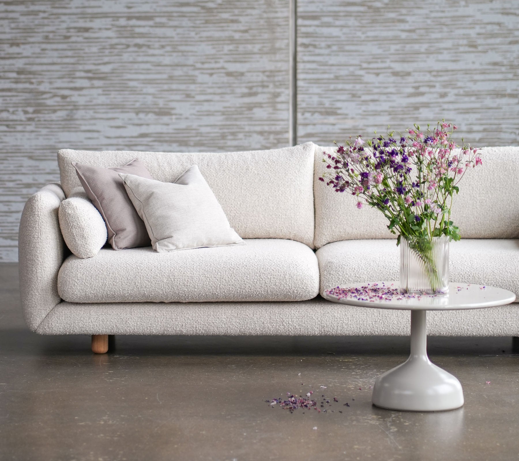 Lys sofa med teakben, pyntet med puter, supplert med et rundt bord og en vase med friske blomster.