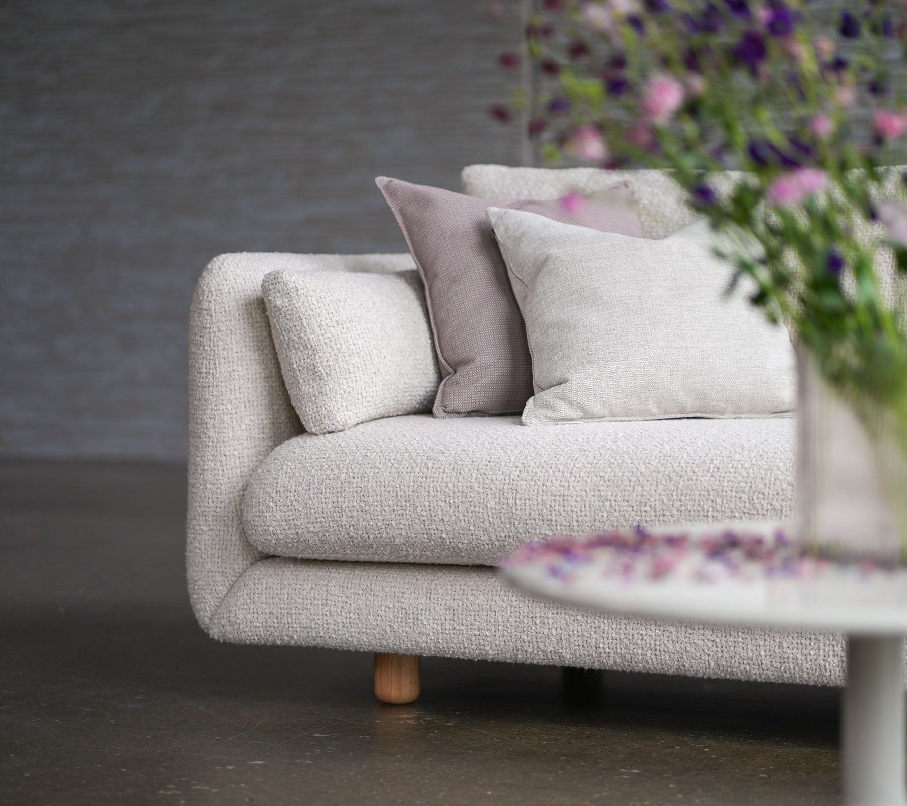 Myk sofa med sandfarget trekk, dekorative puter, teakben og en vas med blomster, som viser Cane-line stil og eleganse.