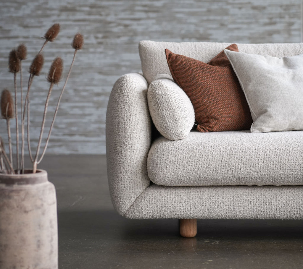 Koselig sofa med blomsterstoff, teakben og opplyftede puter, akkompagnert av dekorativ vase og tørkede blomster. Elegant og innbydende design.