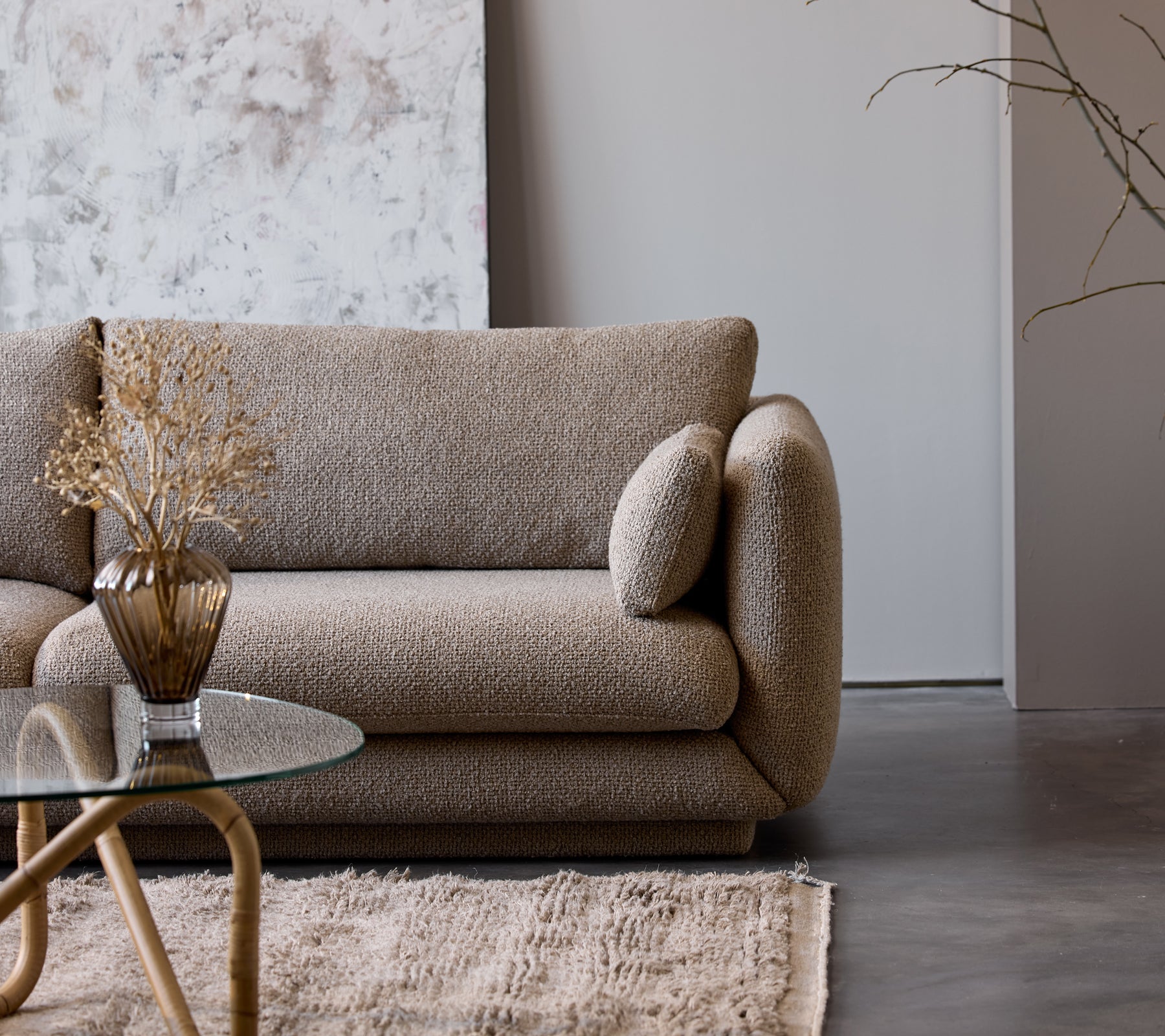 Et taupefarget kaffebord med glassplate står ved siden av en teksturert beige sofa prydet med en dekorativ vase og tørkede blomster.