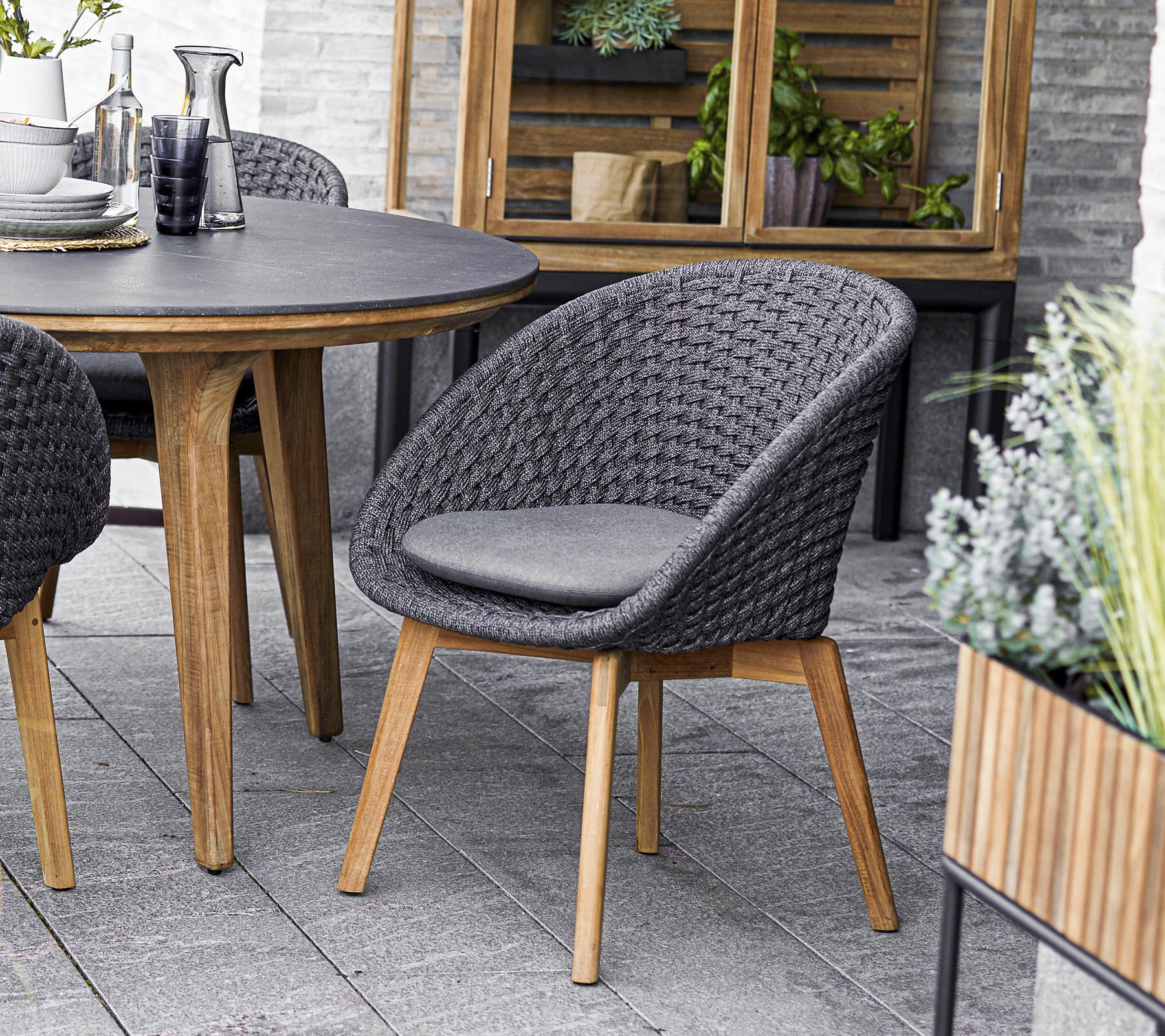 Stilig vevd stol med polstret sete, sammen med et rundt bord og omgitt av grønne planter i krukker. Perfekt for sosiale sammenkomster.