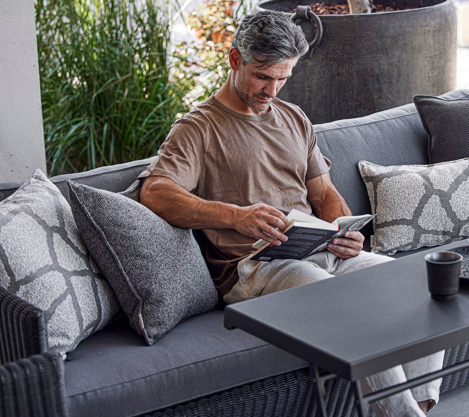 En mann leser en bok mens han sitter på en koselig sofa pyntet med dekorative puter, med grønne planter i bakgrunnen.