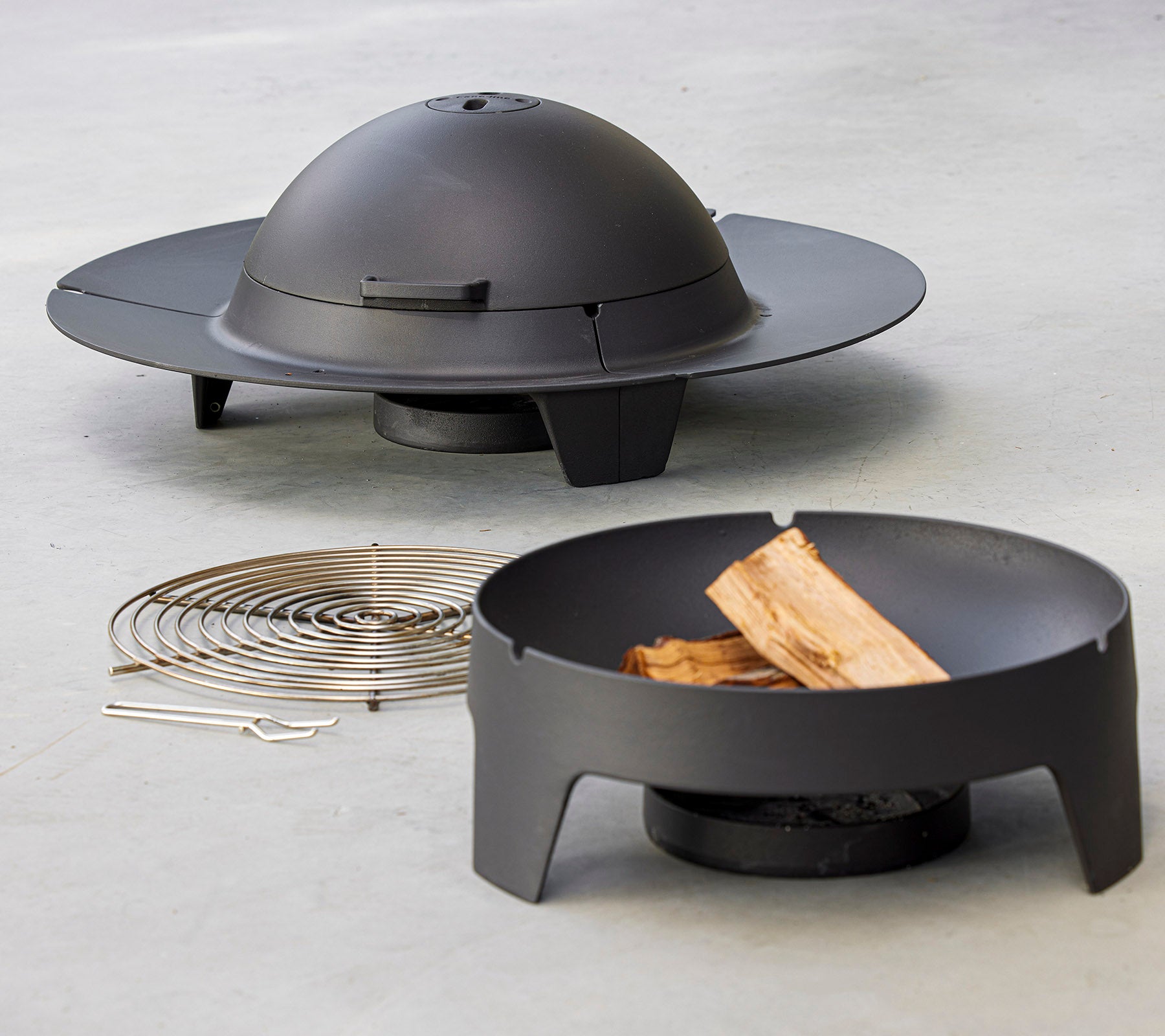 Et moderne bålsted-set med et slankt, rundt design med et kuppellokk, ledsaget av et grillrist og vedstykker.