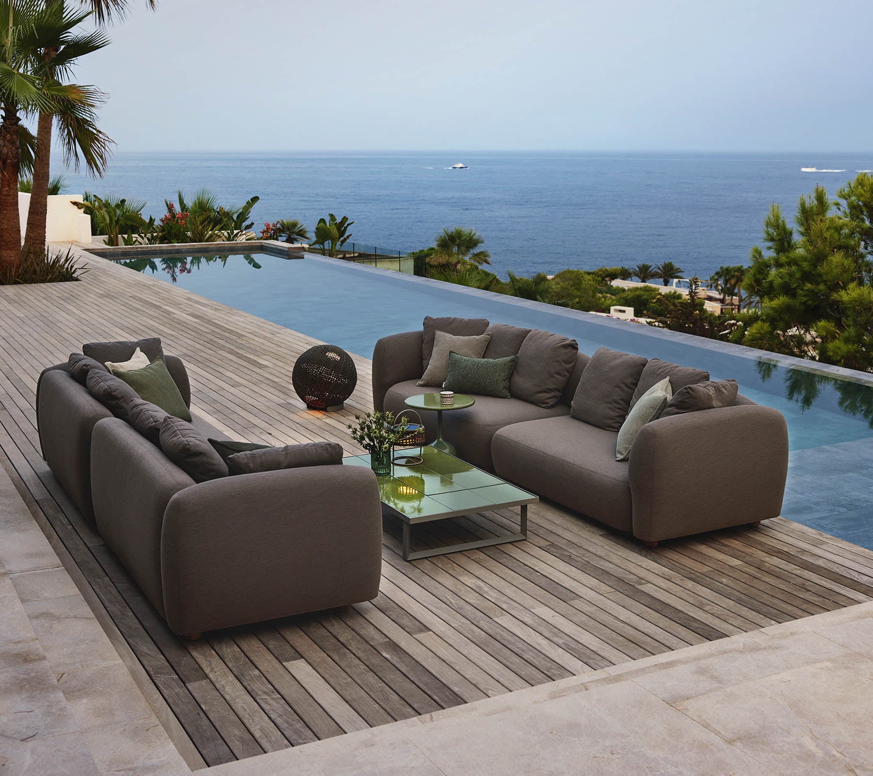 Moderne lounge-møbler ordnet rundt et glasskaffebord, med utsikt over en rolig pool og en utstrakt havutsikt, omgitt av grønt.
