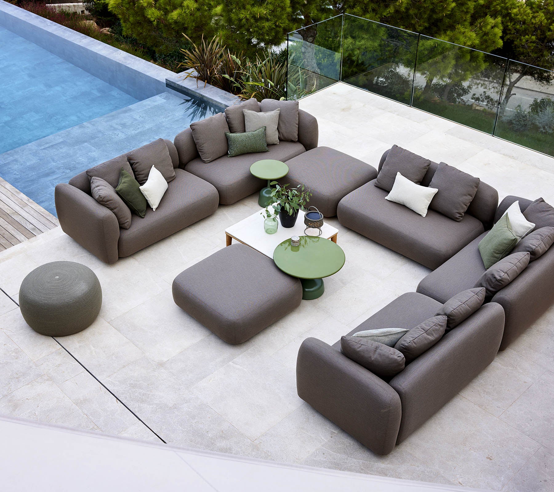 En moderne loungeoppsett med en seksjonalsofa, rund ottoman og grønne aksentbord, omgitt av vegetasjon og en fredelig pool.