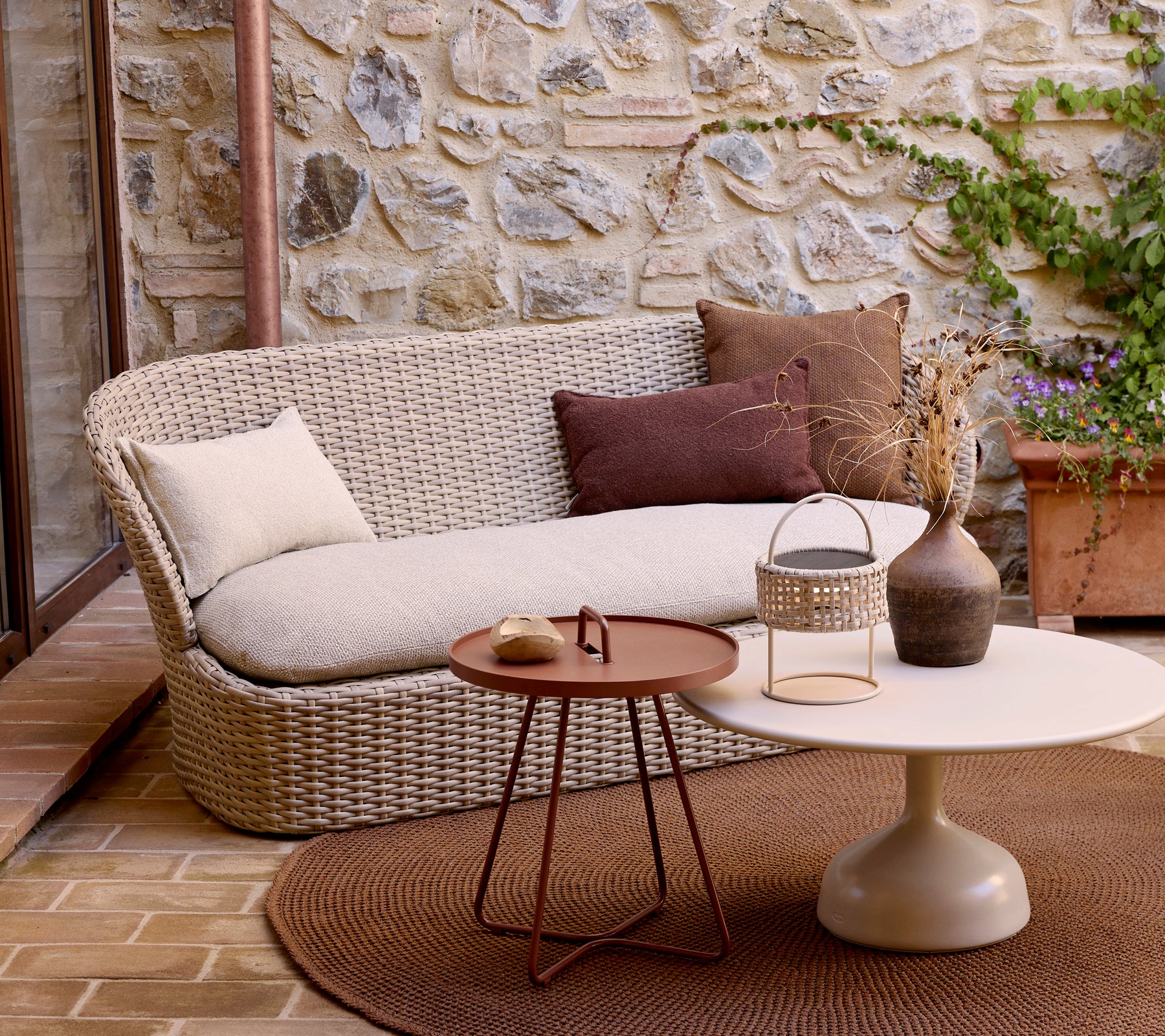 En moderne sofa for to personer med dekorative puter, parret med et rundt kaffebord og planter, stilt opp mot en steinvegg.