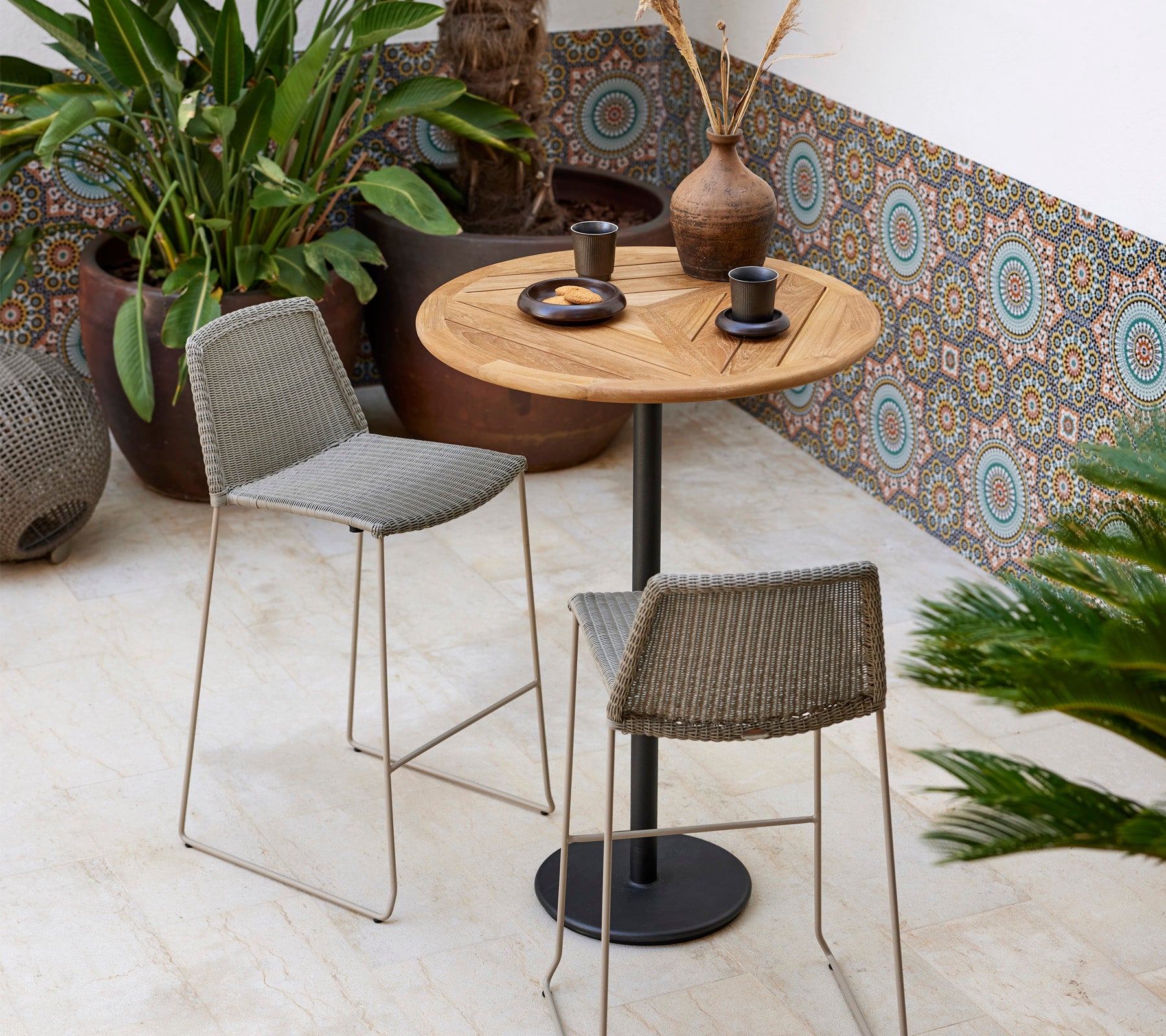 Et stilfullt bistrosett med et rundt treglassbord og to vevde krakker, akkompagnert av dekorative planter og moderne servise.