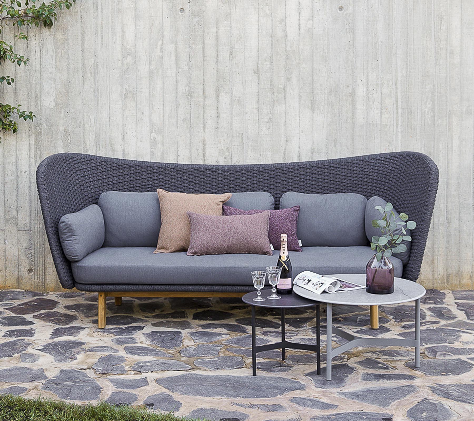 En moderne sofa i grå med dekorative puter, akkompagnert av et lite bord med drikke og en vase, satt mot en teksturert vegg.