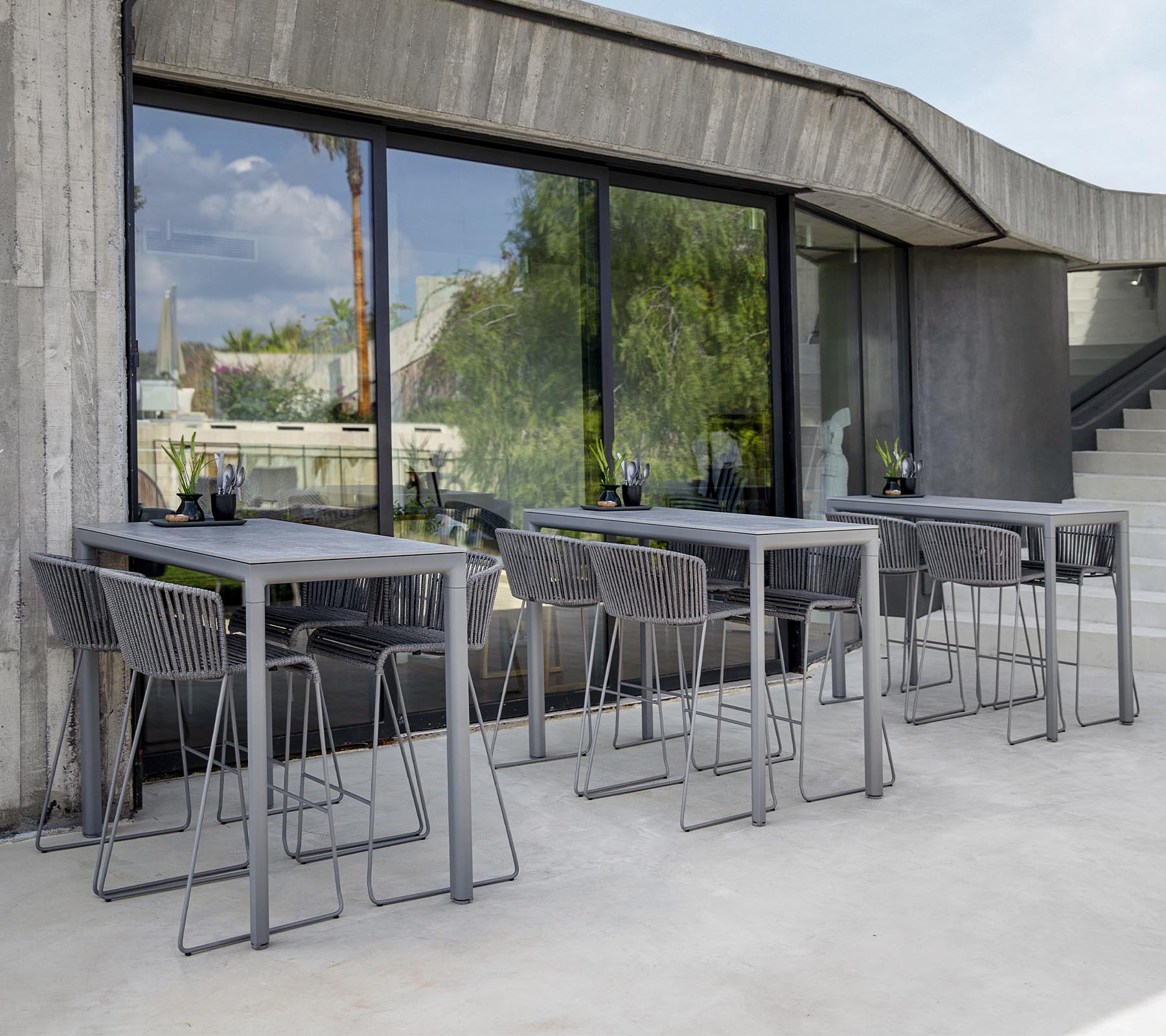 Moderne sitteområde med grå bord og stoler, med store vinduer som reflekterer grønnskapen utenfor. Minimalistisk design estetikk.