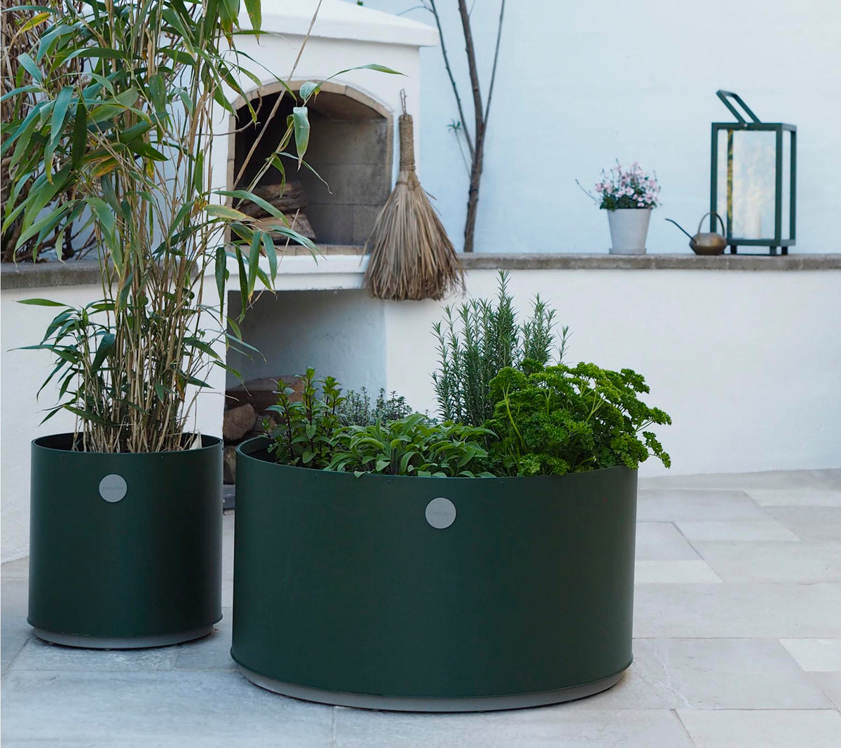 To runde plantekasser i mørkegrønn, den ene med høye planter og den andre fylt med forskjellige urter, står på en lys steinoverflate.