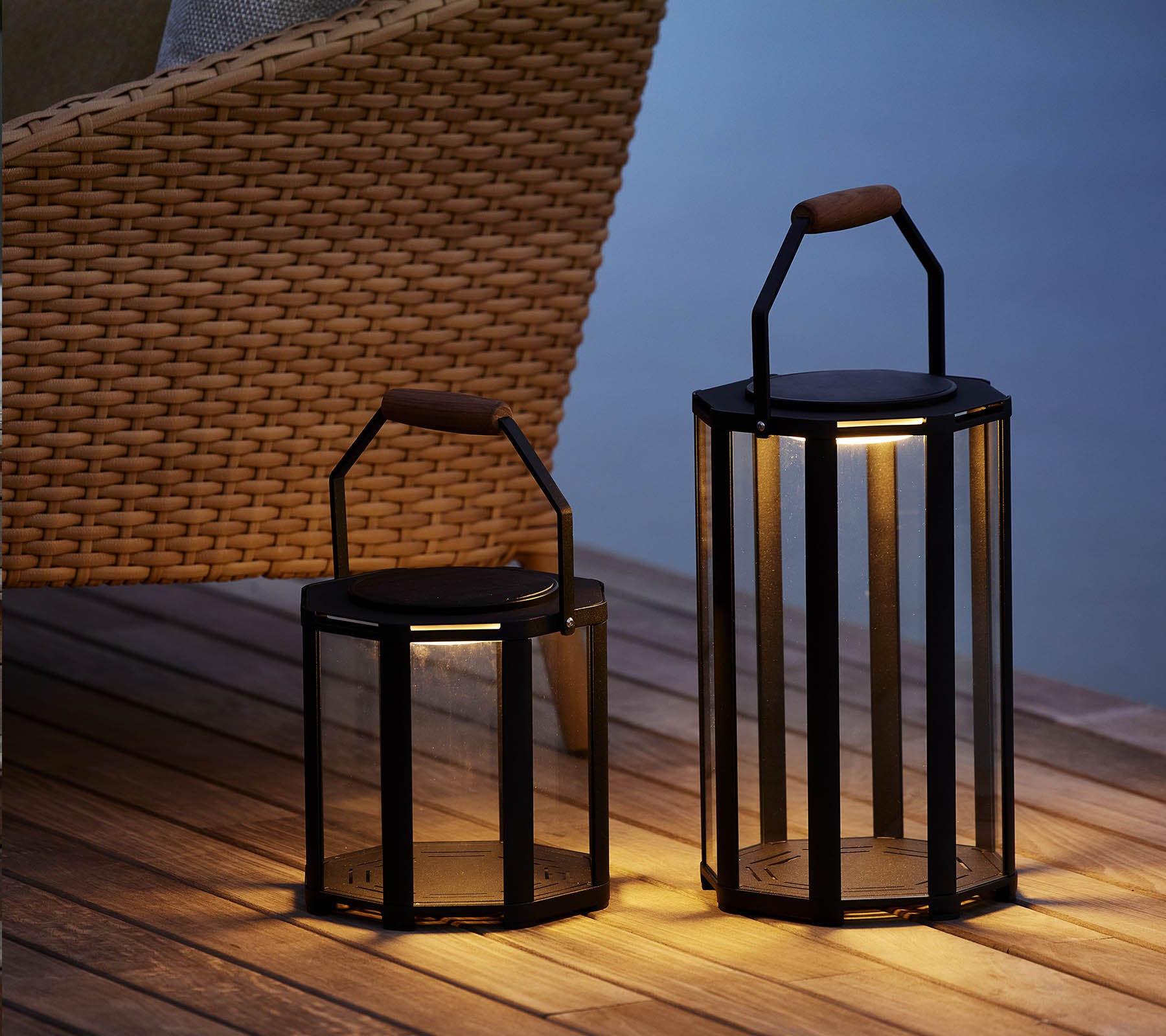 To stilige Lightluxe-lanterner med glasspanser og håndtak, som gir et varmt skjær, plassert ved siden av en vevd stol.