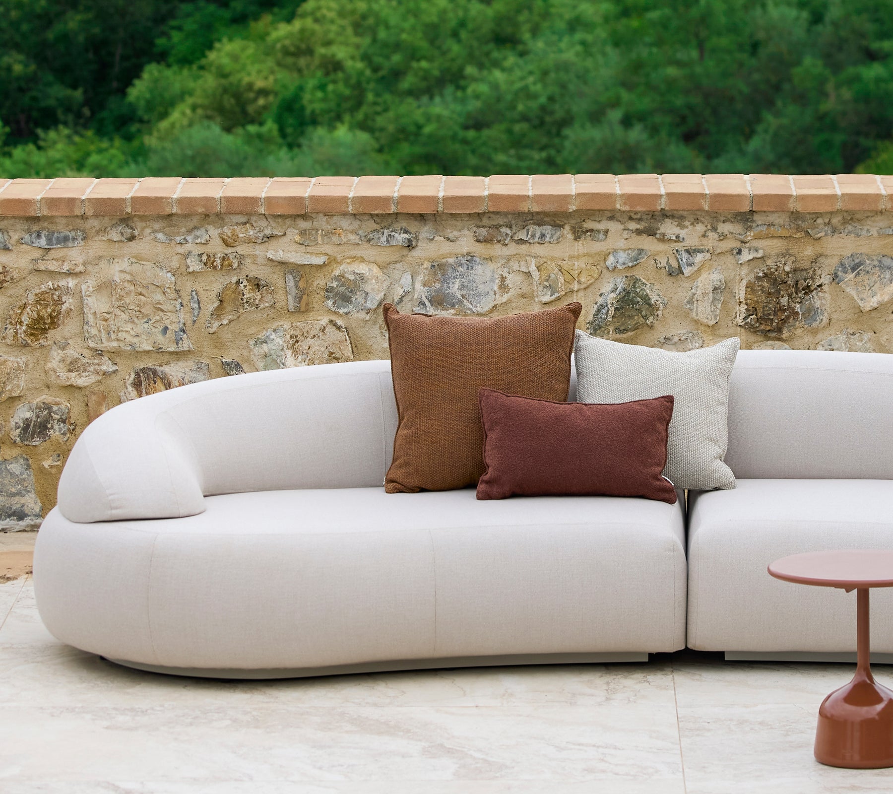 En buet sofa pyntet med Rise-puter i jordfarger ved siden av et stilfullt Glaze-sidebord, mot en steinvegg.