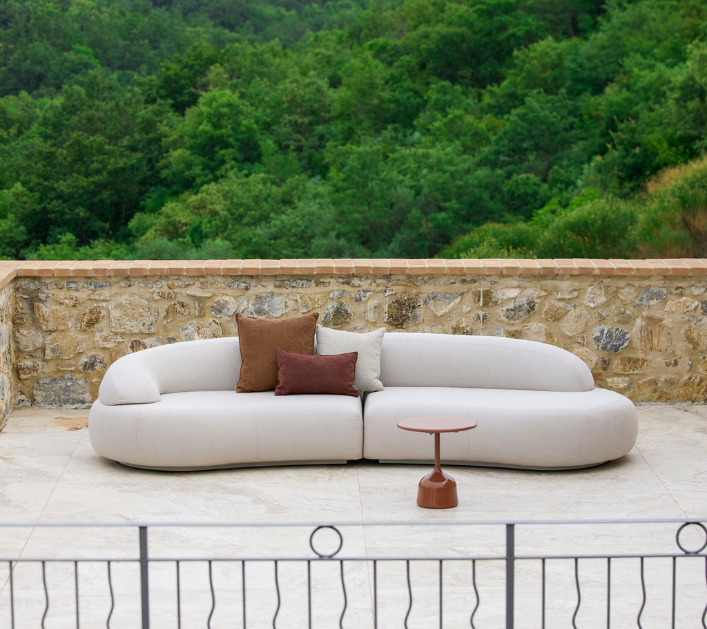 En moderne, buet sofa med nøytral trekk og dekorativeputer, supplert med et stilrent sidebord i en naturlig setting.