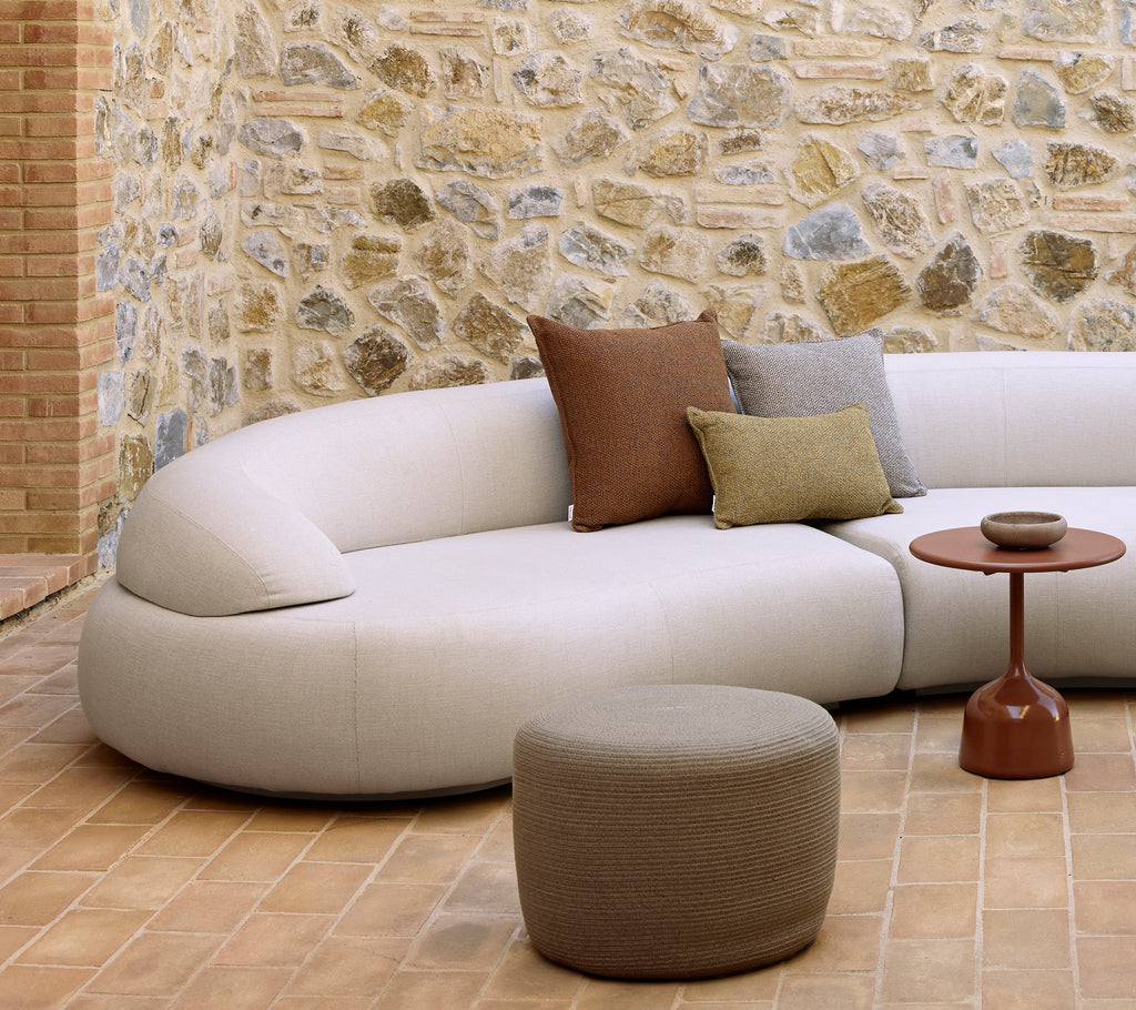 Bøyd sofa med dekorative puter, et sirkulært fotteppe og et glasert sidebord på et teksturert gulv med en steinvegg i bakgrunnen.