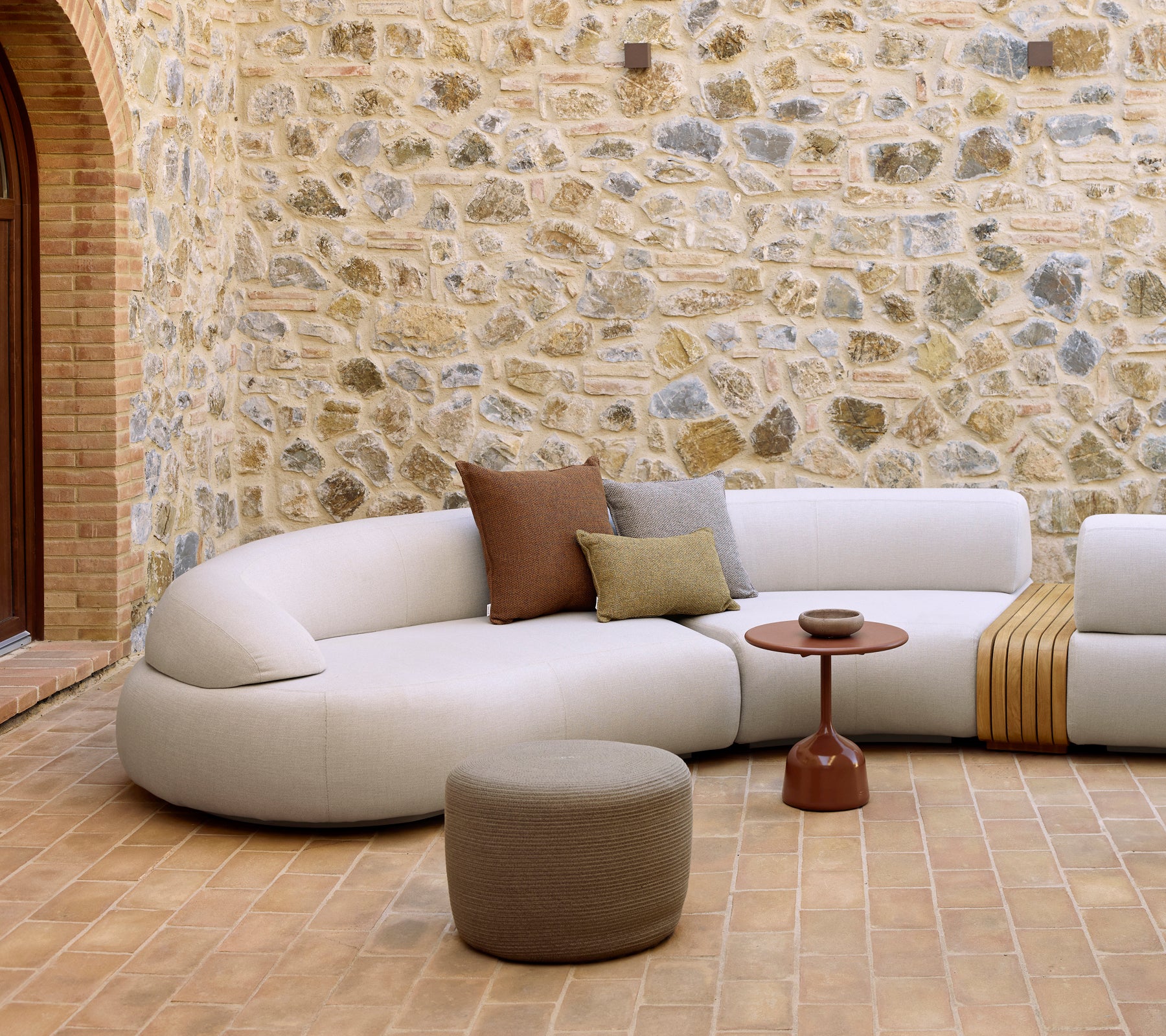 Behagelig loungeområde med en buet sofa, Glaze kaffebord og en rund fotpall mot en teksturert steinvegg.
