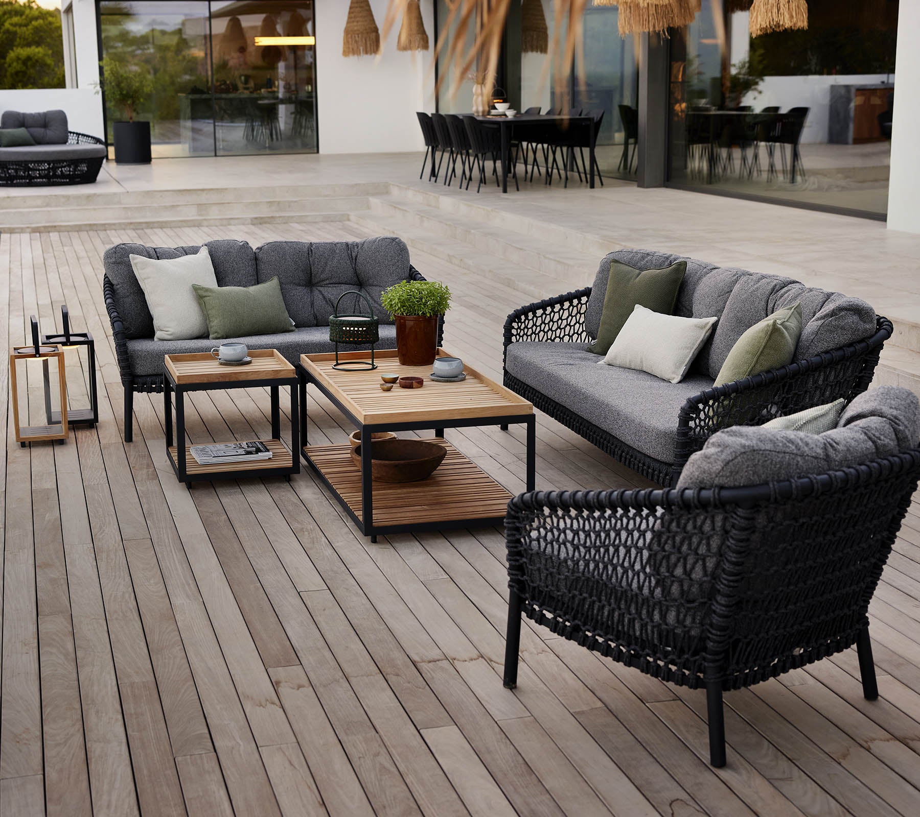 Stilfullt utendørs loungeområde med svart flettmøbler, grå puter, et bord i tre og potteplanter på en treterrasse.