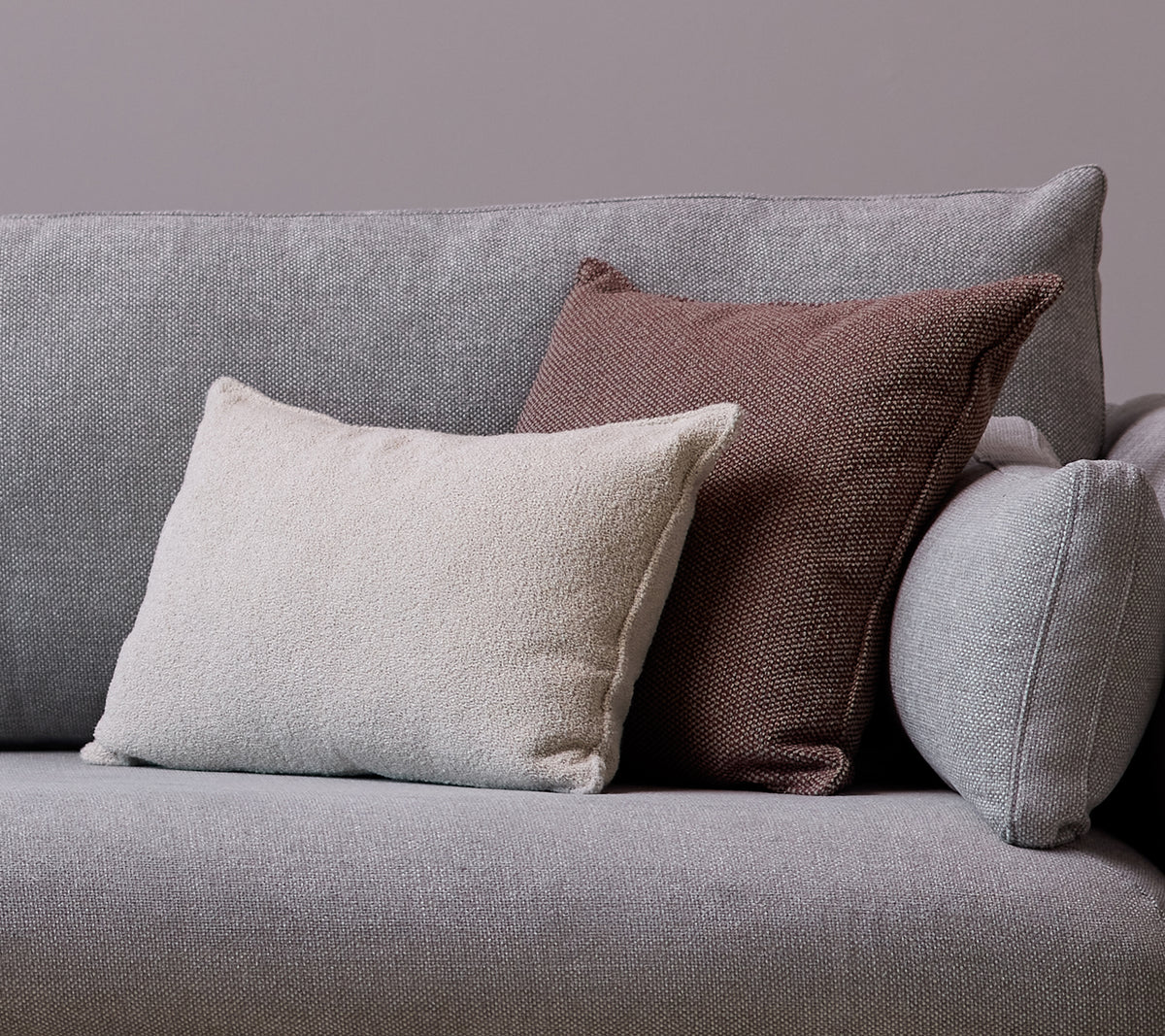 En hvit Scent scatterpute og en Wove mørk bordeaux pute er plassert på en grå sofa.
