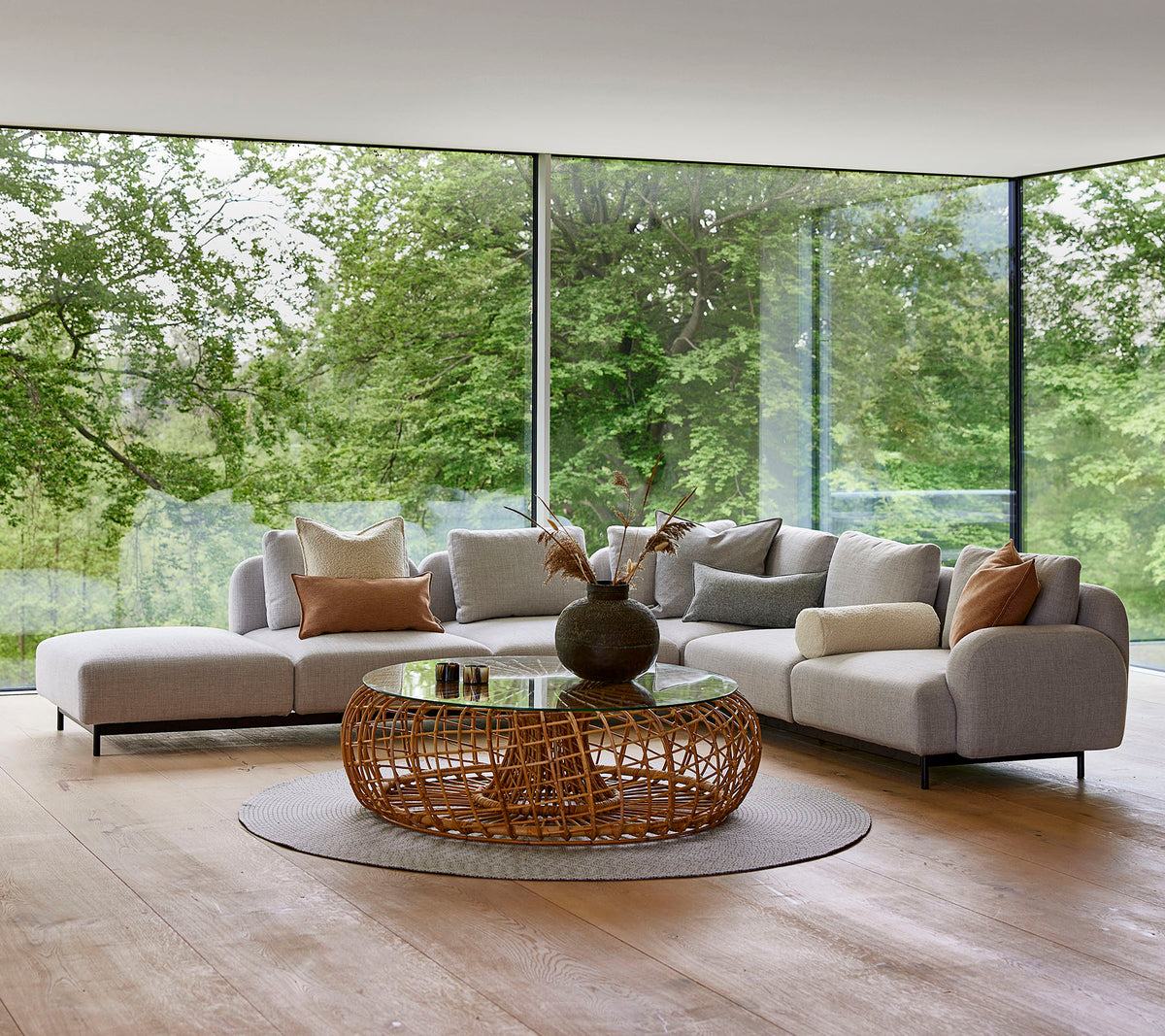 Moderne stueområde med en modulsofa med dekorative puter, et rundt, vevd kaffebord og store glassvinduer som viser grøntområder.