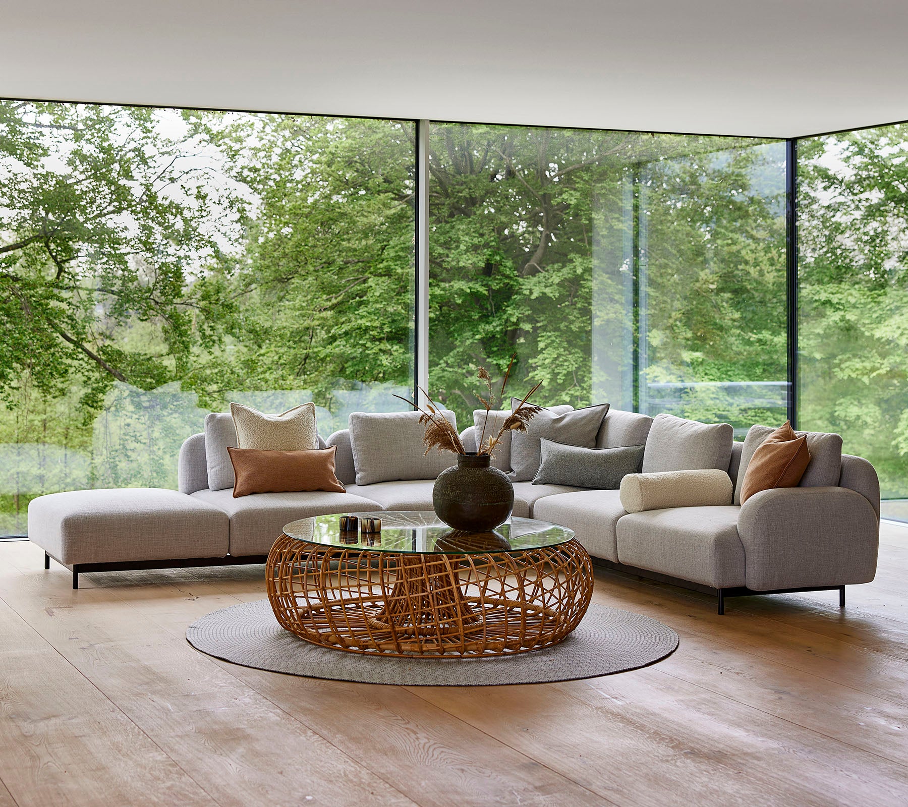 Moderne stueområde med en modulsofa med dekorative puter, et rundt, vevd kaffebord og store glassvinduer som viser grøntområder.