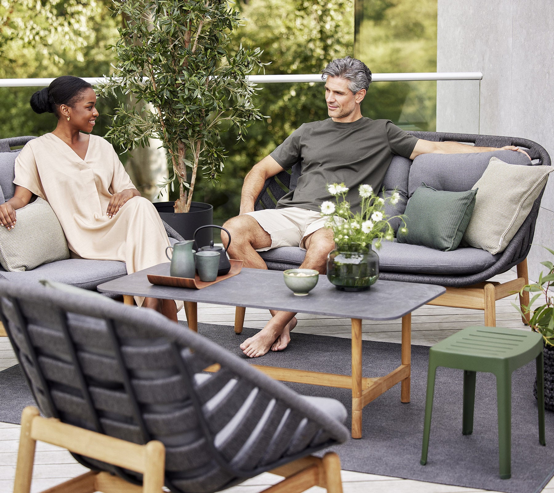 To personer slapper av på en stilfull utendørs sofa, omgitt av grønt og dekorative planter, og nyter et fredelig øyeblikk sammen.